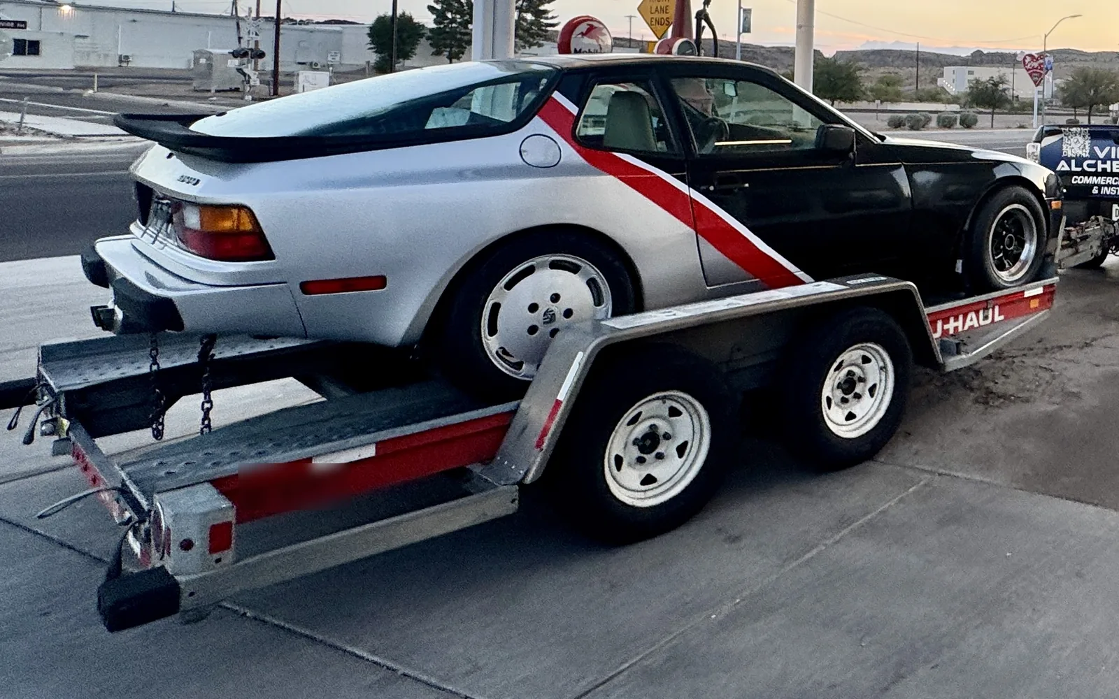 Porsche 944 in a silver and black two-tone with a red stripe accent, loaded on a trailer